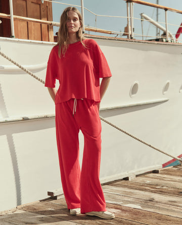 A person stands on a dock beside a white boat, wearing THE GREAT. SU26 The Micro Terry Track Crew in Pimento with matching pants and white shoes.