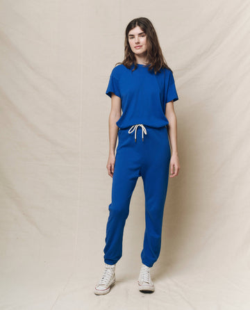 A person stands against a beige backdrop, exuding a relaxed style with THE GREAT. SP25 KNITS' The Boxy Crew in Bright Cobalt and joggers, complemented by white sneakers for an effortlessly cool look.