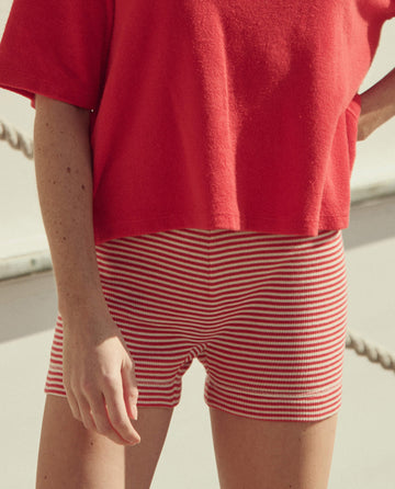 Wearing THE GREAT. SU26 Relay Short in Pimento Stripe, the person stands outdoors with parts of their arm and legs visible.