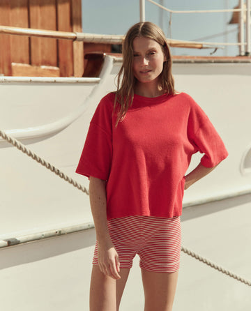 A woman stands outdoors in front of a white boat, wearing a red short-sleeve top and THE GREAT. SU26's The Relay Short in Pimento Stripe.