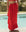 Wearing THE GREAT. SP25 SWIM’s The Pareo. -- Hot Red, this person stands poolside amid lush greenery, showcasing perfect beachwear with a bright red ruffled sarong skirt and matching top.