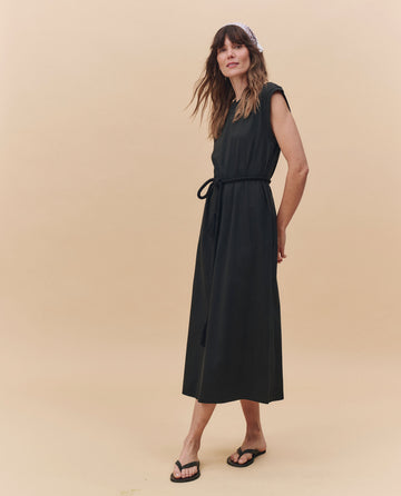 A woman wears THE GREAT. PF25 HEAVY JERSEY KNITS The Square Tee Dress in Almost Black, a sleeveless ankle-length jersey dress with waist tie, styled with black sandals and a light headscarf, standing on a beige background.