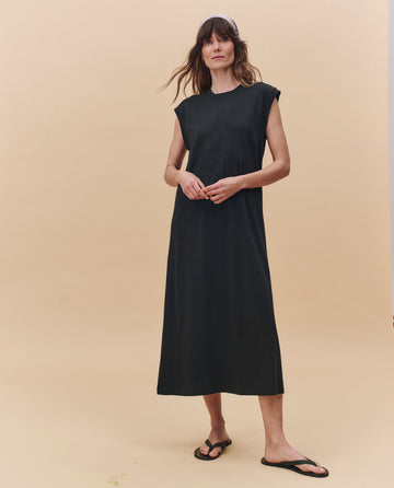 A woman stands against a beige background wearing THE GREAT. PF25 HEAVY JERSEY KNITS The Square Tee Dress in Almost Black, paired with black sandals.