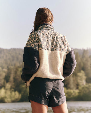 A person in The Plush Terrain Half-Zip jacket, Falling Daisy Colorblock by THE GREAT. SP25 TGO, stands outdoors near plush terrain and a body of water, facing away.