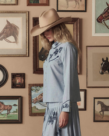A person in THE GREAT. STETSON SP26 Chambray Embroidered Western Shirt and a wide-brimmed tan hat stands before a wall with framed horse art.