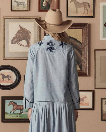 Wearing THE GREAT. STETSON SP26 Chambray Embroidered Western Shirt, a person in a cowboy hat stands facing a wall adorned with framed horse paintings and photographs.