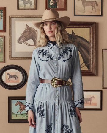 A woman in a light blue dress and a beige cowboy hat stands before framed horse paintings, wearing THE GREAT. STETSON SP26's The Big Buckle Belt in Cognac With Brass.