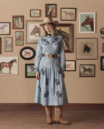 A woman wears THE GREAT. STETSON SP26's The Big Buckle Belt in Cognac With Brass, paired with a light blue western dress, cowboy hat, and boots, standing before a wall adorned with framed horse art.