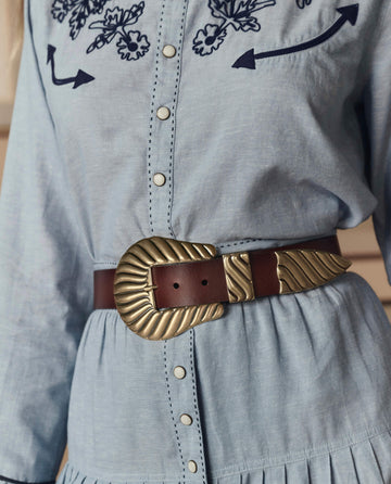 Close-up of a person wearing a light blue, embroidered dress, paired with THE GREAT. STETSON SP26 The Big Buckle Belt in cognac with a large, textured brass buckle.