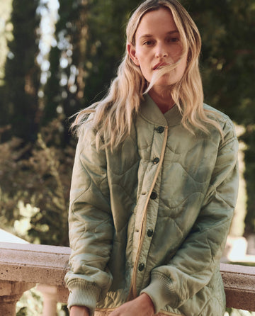 A person with long hair stands outdoors, wearing The Quilted Liner Jacket in mottled green by THE GREAT. SP25 D1, framed by a serene backdrop of trees.