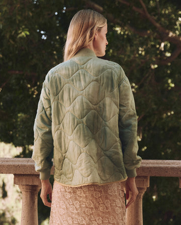 A blonde-haired individual, back turned, leans on a stone railing outdoors. They're wearing a Mottled Green "The Quilted Liner Jacket" by THE GREAT. SP25 D1 and a lace skirt, with trees in the background.