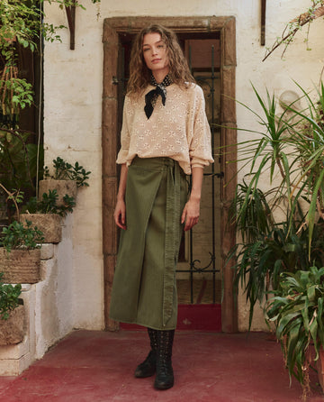 A person stands in front of a doorway with potted plants, wearing a light patterned blouse, green wrap skirt, black boots and neck scarf, and The Crochet Pullover in Natural by THE GREAT. PF25.