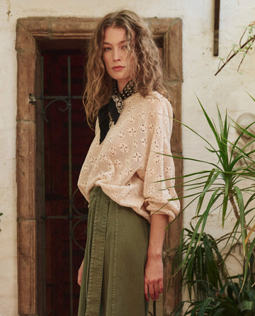 A woman with wavy hair wears THE GREAT. PF25 The Crochet Pullover – Natural, paired with a green skirt and black scarf, standing indoors by plants and a rustic doorway.