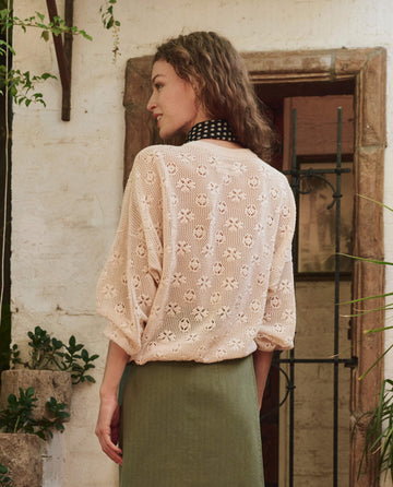 A woman stands with her back to the camera, wearing THE GREAT. PF25 The Crochet Pullover in Natural and a green skirt, set against a rustic backdrop with plants and a white textured wall.