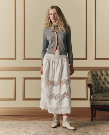 A woman stands indoors wearing The Dainty Cardigan in Heather Grey by THE GREAT. SP26 D2, paired with a long white lace skirt and beige lace-up boots, while a vintage-style sofa sits in the background.