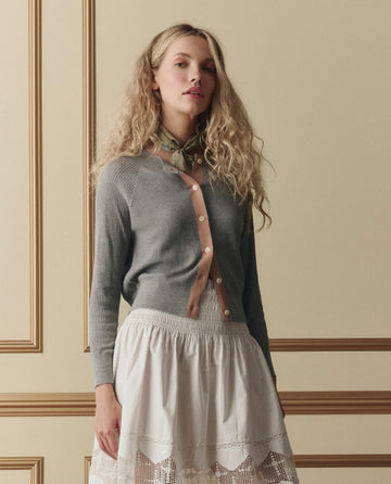 A woman with long, wavy blonde hair wears THE GREAT. SP26 D2's The Dainty Cardigan in heather grey with pink buttons, plus a white skirt and patterned neck scarf, standing before a beige paneled wall.