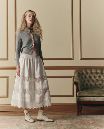 A woman stands indoors next to a green upholstered sofa, wearing THE GREAT. SP26 D2's The Rosette Crochet Canary Skirt in cream, paired with a gray cardigan, light scarf, and lace-up boots.
