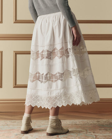 Indoors, a person wears a gray top, beige ankle boots, and THE GREAT. SP26 D2's The Rosette Crochet Canary Skirt in cream—a long white crochet skirt with lace panels and scalloped hem.