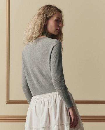 A woman with wavy blonde hair faces sideways against a beige wall with molding, wearing THE GREAT. SP26 D2's The Dainty Cardigan in Heather Grey and a white skirt.
