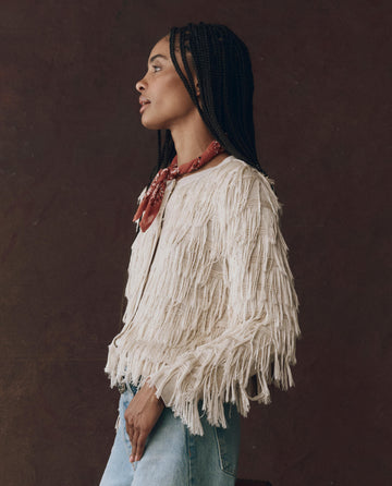 A woman in profile wears THE GREAT. F25 D1 The Fringe Cardigan in Natural, styled with a red bandana and light blue jeans, standing against a dark background.