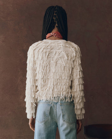A person stands with their back to the camera, wearing THE GREAT. F25 D1 The Fringe Cardigan in Natural—crafted from viscose yarn—paired with blue jeans and a red bandana, set against a plain brown background.
