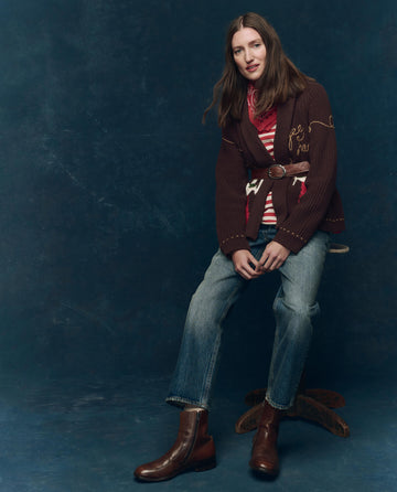 A woman with long brown hair sits on a wooden stool against a blue background, wearing a brown jacket, striped shirt, and THE GREAT. SP25 COLLECTORS The Wayne Jean in Montana Wash with brown boots.