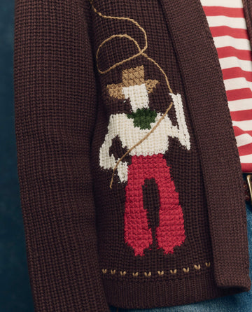 Close-up of THE GREAT. F25 COLLECTORS The Lasso Lodge Cardigan in umber, crafted from a soft cotton blend with stitched cowboy and lasso details, layered over a red and white striped shirt.