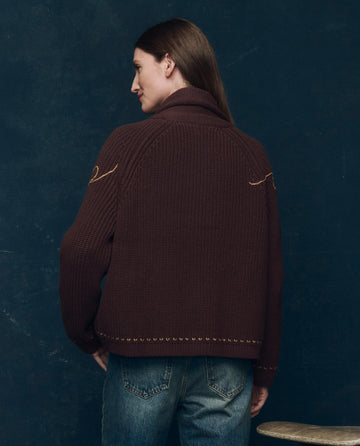 A person with long brown hair faces away, wearing THE GREAT. F25 COLLECTORS The Lasso Lodge Cardigan in Umber and blue jeans, standing against a dark blue background.