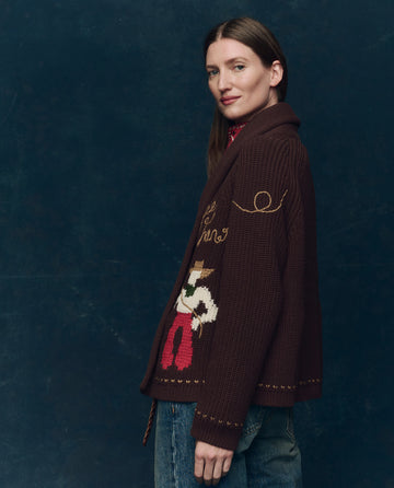 A woman with straight brown hair wears THE GREAT. F25 COLLECTORS' The Lasso Lodge Cardigan in Umber and blue jeans, standing before a dark blue background and looking sideways at the camera.