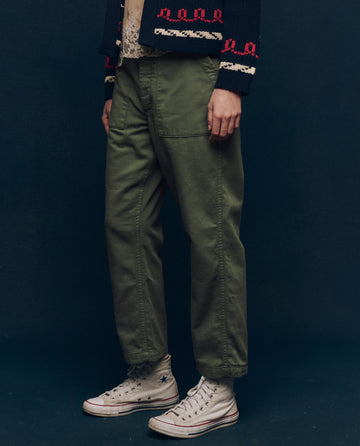 Someone wearing The Vintage Army Pant by THE GREAT. SP25 COLLECTORS, a dark jacket with designs, and white high-top sneakers stands against a dark backdrop.