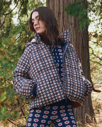 A woman stands outdoors in front of trees, wearing THE GREAT. F25 TGO The Whistler Puffer – Mini Frosted Daisy, a layering jacket with recycled insulation, and matching floral pants.