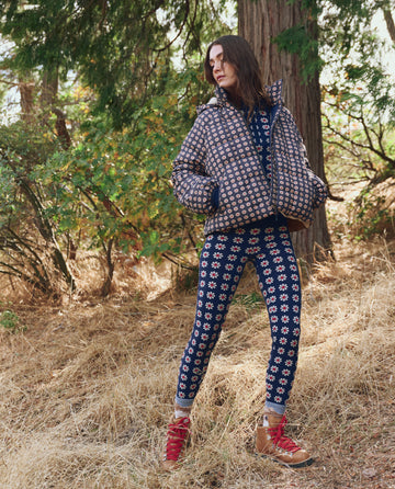 A person stands outdoors wearing THE GREAT. F25 TGO's The Whistler Puffer in Mini Frosted Daisy with matching pants, featuring recycled insulation, paired with red hiking boots on dry grass among trees and greenery.