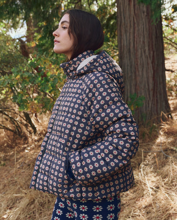 A person wears THE GREAT. F25 TGO’s The Whistler Puffer in Mini Frosted Daisy outdoors among trees and dry grass. The jacket’s recycled insulation provides warmth and sustainable style.