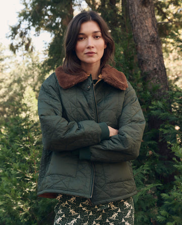 Woman stands outdoors in front of trees, wearing THE GREAT. F26 TGO The Reversible Logger Puffer. -- Pine And Amber, arms crossed and looking at the camera.