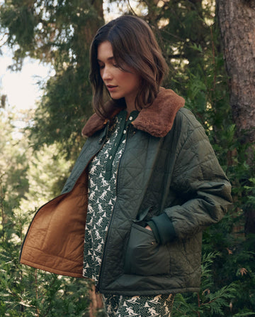 A woman stands outdoors among trees, wearing THE GREAT. F26 TGO The Reversible Logger Puffer. -- Pine And Amber over a patterned dress.