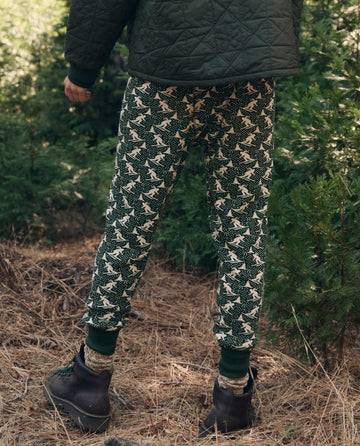Person wearing The Union Long John in Spruce dark green with a repeating white skier pattern, a dark quilted jacket, and brown hiking boots.