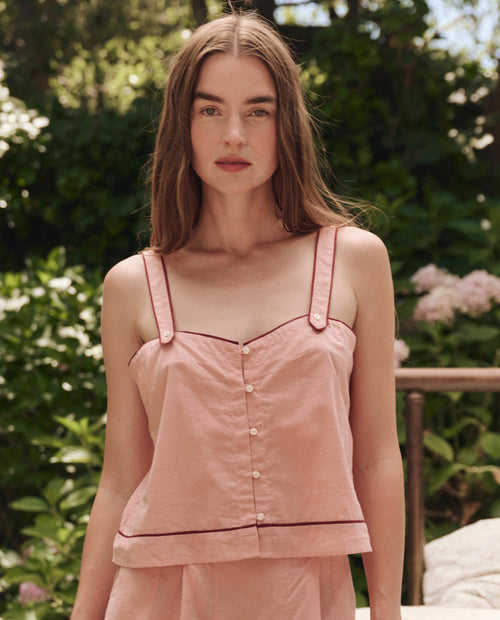 A woman with long brown hair wears THE GREAT. SP25 SLEEP The Western Sleep Cami in Rose Bloom, standing outdoors amid lush green garden foliage.