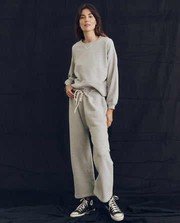 A person in "The League Sweatshirt" by THE GREAT. PS24 Lofty Knits in Lofty Heather Grey and matching sweatpants, standing against a dark black background. They are wearing black and white sneakers, handcrafted locally in Los Angeles.