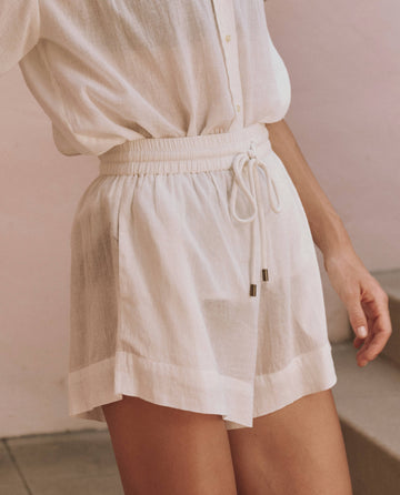 A person wearing a matching light-colored, loose-fitting shirt and THE GREAT. SP25 SWIM The Delta Short in white, made of lightweight fabric, standing indoors near a staircase.
