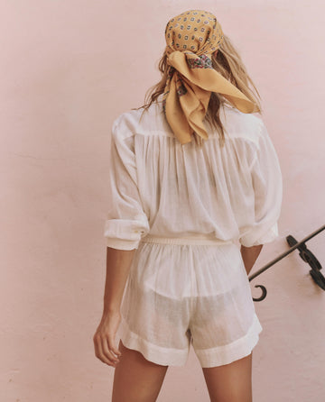 Person standing with their back to the camera, wearing a white long-sleeve shirt, THE GREAT. SP25 SWIM The Delta Short in White, and a yellow patterned headscarf against a light pink wall.