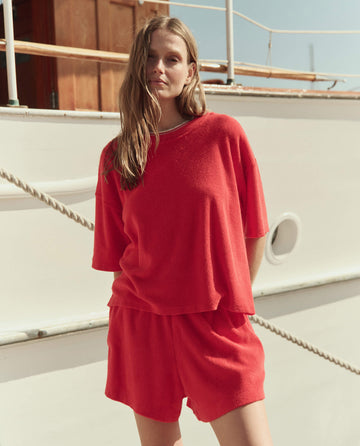 A woman stands by a boat, wearing THE GREAT. SU26 The Micro Terry Track Crew in Pimento with matching red shorts.
