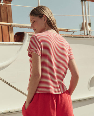 A person with light brown hair stands outdoors beside a white boat, wearing THE GREAT. SU26’s The Little Tee -- Pimento Stripe and red shorts.