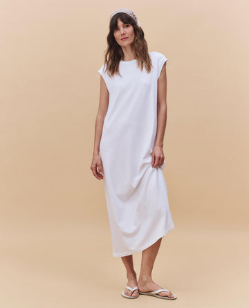 Against a beige background, a woman wears THE GREAT. PF25 HEAVY JERSEY KNITS The Square Tee Dress in True White, featuring a boxy fit, paired with a headband and white flip-flops.