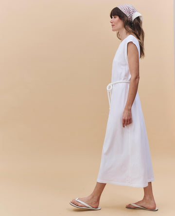 A woman wears THE GREAT. PF25 HEAVY JERSEY KNITS The Square Tee Dress in True White, sleeveless and boxy, styled with a rope belt, white sandals, and a white headscarf against a beige background.