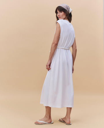 A woman, facing away, wears THE GREAT. PF25 HEAVY JERSEY KNITS The Square Tee Dress in True White—sleeveless and boxy—paired with a beige headscarf and white sandals, set against a plain beige background.