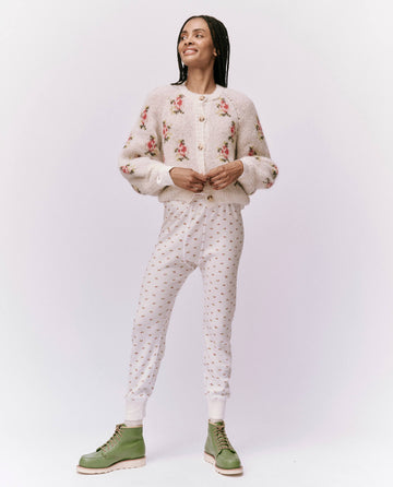 A person wearing The Waffle Long John in White Mini Heirloom Rose from THE GREAT. HOL 24 ROSEBUD CAPSULE stands against a white background, smiling warmly in their cozy attire, complemented by patterned pants and green boots.