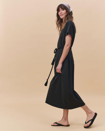 Woman wearing THE GREAT. PF25 HEAVY JERSEY KNITS The Boxy Tee Dress in Almost Black, styled with a cotton rope belt, black flip-flops, and a patterned headscarf, standing against a beige background.