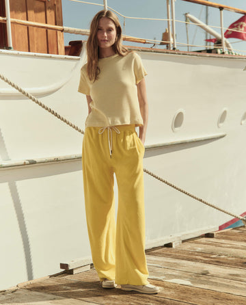 Woman stands on a dock before a white boat, wearing THE GREAT. SU26's The Little Tee in Soft Yellow Stripe, loose yellow pants, and beige shoes.