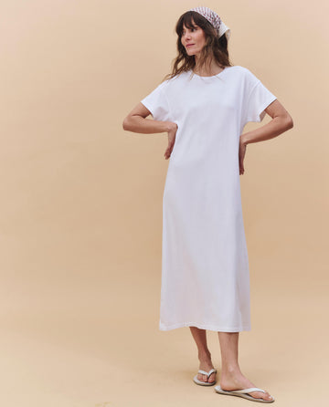 A woman stands against a beige background wearing THE GREAT. PF25 HEAVY JERSEY KNITS The Boxy Tee Dress in True White, styled with a white patterned headscarf and white flip-flops.