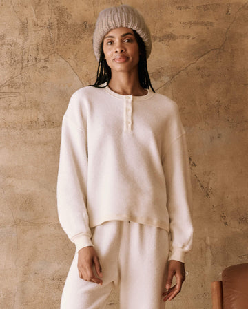 A woman wears The Handknit Chunky Beanie in cream, by THE GREAT. HOL 24 PLUSH KNITS, enhancing her cream sweater and matching pants as she poses elegantly against a textured beige wall.